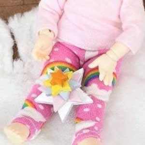 May include: A doll with blonde hair wearing a pink shirt and rainbow leggings sits on a white fluffy rug. The doll is holding a colorful star-shaped toy.
