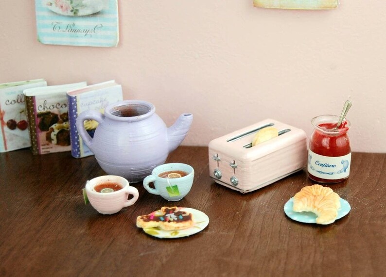Pu&ograve; includere: Un set da cucina in miniatura con una teiera lavanda, due tazzine da t&egrave; rosa, un tostapane rosa, un piatto con un dolce, un piatto con un croissant e un vasetto di marmellata rossa etichettato "Confiture".