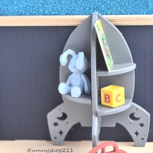 May include: A gray rocket-shaped bookshelf with three shelves. The top shelf has a book, the middle shelf has a blue toy, and the bottom shelf has a yellow block with the letters "B" and "C". A red toy car is in front of the bookshelf.