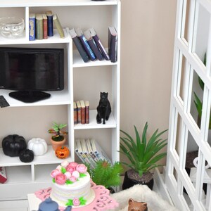 Pu&ograve; includere: Una libreria bianca con una statuetta di gatto nero, libri e una pianta in vaso. Un televisore nero &egrave; sullo scaffale superiore. Un letto per cani bianco con una statuetta di cane marrone &egrave; sul pavimento. Una torta rosa con glassa bianca e fiori rosa &egrave; su un supporto per torta rosa. Un bollitore blu e tazze da t&egrave; sono su un tavolo bianco.
