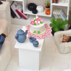 Pu&ograve; includere: Una torta bianca con glassa rosa e fiori su un supporto per torte rosa e bianco. C'&egrave; una teiera blu e due tazze da t&egrave; blu su un tavolo bianco. La torta &egrave; in un ambiente di casa delle bambole.
