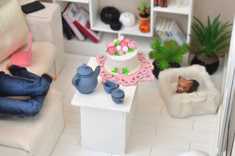 Pu&ograve; includere: Una torta bianca con glassa rosa e fiori su un supporto per torte rosa e bianco. C'&egrave; una teiera blu e due tazze da t&egrave; blu su un tavolo bianco. La torta &egrave; in un ambiente di casa delle bambole.