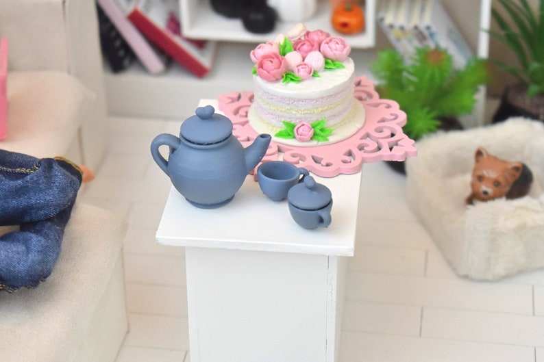 Pu&ograve; includere: Un set da t&egrave; in miniatura con una teiera blu, due tazze e una zuccheriera. Il set da t&egrave; &egrave; su un tavolo bianco accanto a una torta rosa decorata con fiori rosa.