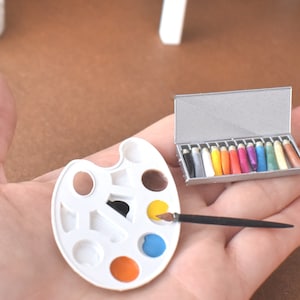 May include: A miniature artist's palette with a paintbrush and a closed box of miniature paint tubes. The palette has eight wells, some filled with paint. The paint tubes are arranged in a row and are various colours.