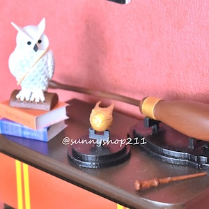 May include: A white owl figurine perched on a stack of books, a golden snitch on a black stand, a brown broomstick on a black stand, and a brown wooden wand on a wooden surface with red and yellow stripes.
