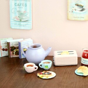 Pu&ograve; includere: Un set da cucina in miniatura con una teiera viola, due tazze da t&egrave;, un tostapane rosa, un barattolo di marmellata e un croissant su un piatto blu. Il set &egrave; su un tavolo di legno con tre libri di cucina in miniatura.