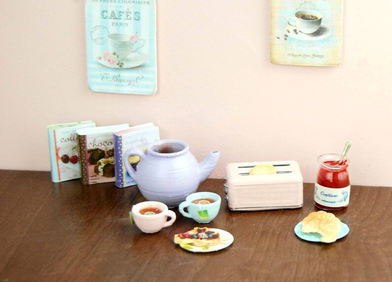 Pu&ograve; includere: Un set da cucina in miniatura con una teiera viola, due tazze da t&egrave;, un tostapane rosa, un barattolo di marmellata e un croissant su un piatto blu. Il set &egrave; su un tavolo di legno con tre libri di cucina in miniatura.