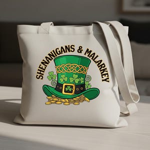 May include: A cream-colored tote bag featuring a green leprechaun hat design with gold coins. The hat has a gold Celtic knot and shamrocks. The words "SHENANIGANS & MALARKEY" are printed in a semi-circle around the hat.