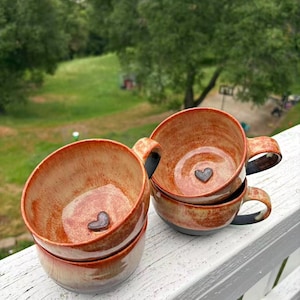 May include: Four handmade ceramic mugs stacked on a white surface. The mugs feature a heart-shaped detail inside and a warm, earthy colour scheme, transitioning from beige to reddish-brown. Each mug has a black handle.