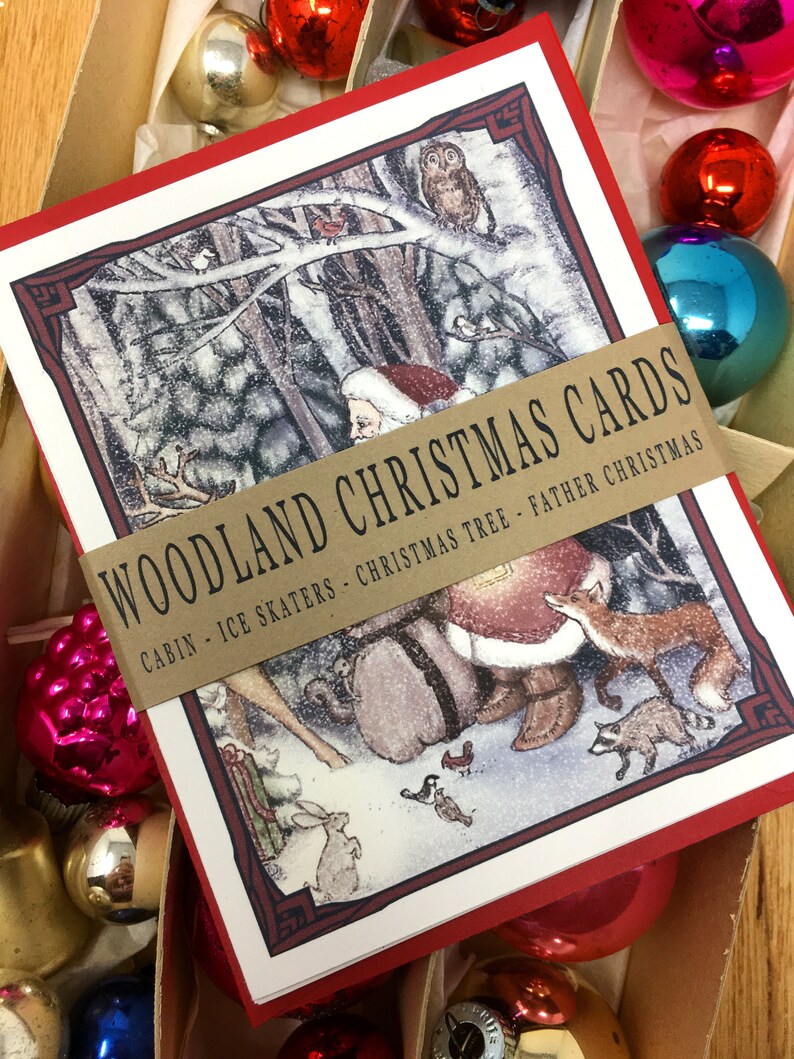 May include: A set of red and white Christmas cards with a woodland scene featuring Santa Claus, a fox, a raccoon, a rabbit, and birds. The cards have a brown ribbon with the text "WOODLAND CHRISTMAS CARDS CABIN - ICE SKATERS - CHRISTMAS TREE - FATHER CHRISTMAS".
