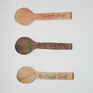 Peut inclure: Trois cuillères en bois avec des étiquettes manuscrites : "Soupe de cerises", "Soupe aux champignons" et "Soupe de courge butternut".