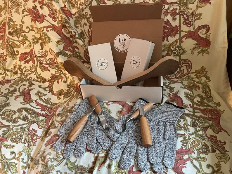K&ouml;nnte beinhalten: Eine Schachtel mit zwei Holzl&ouml;ffeln, zwei Paar grauen Schutzhandschuhen und zwei kleinen Werkzeugen mit Holzgriffen. Die Schachtel ist ge&ouml;ffnet und die Gegenst&auml;nde sind sichtbar. Die Schachtel steht auf einem gemusterten Stoff.