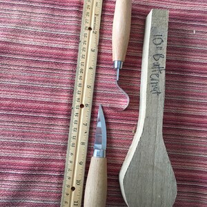 May include: Two wood carving tools with wooden handles and a ruler. The tools are a hook knife and a small knife. The ruler is 12 inches long. The tools are on a red and white striped fabric.