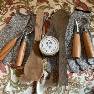 K&ouml;nnte beinhalten: Ein Satz Holzschnitzwerkzeuge, darunter zwei Schnitzmesser, ein Hakenmesser, ein Holzl&ouml;ffel, ein Holzstift und eine Dose Holzschnitzwachs. Die Werkzeuge liegen auf einem floral gemusterten Stoff.
