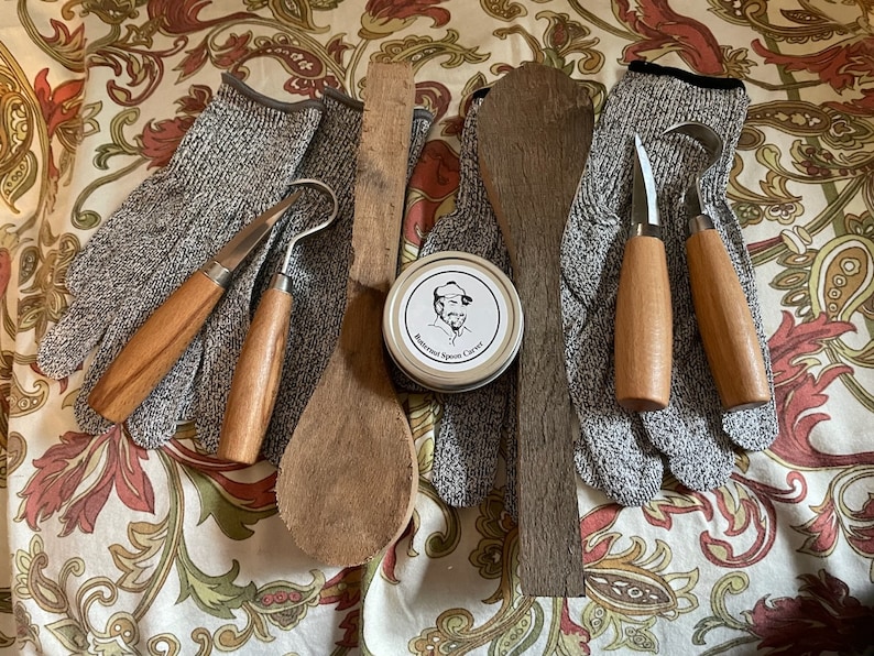 K&ouml;nnte beinhalten: Ein Satz Holzschnitzwerkzeuge, darunter zwei Schnitzmesser, ein Hakenmesser, ein Holzl&ouml;ffel, ein Holzstift und eine Dose Holzschnitzwachs. Die Werkzeuge liegen auf einem floral gemusterten Stoff.