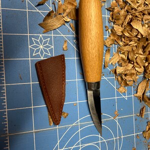 May include: A small, wooden-handled carving knife with a curved blade, resting on a blue cutting mat with wood shavings. A brown leather sheath is next to the knife.