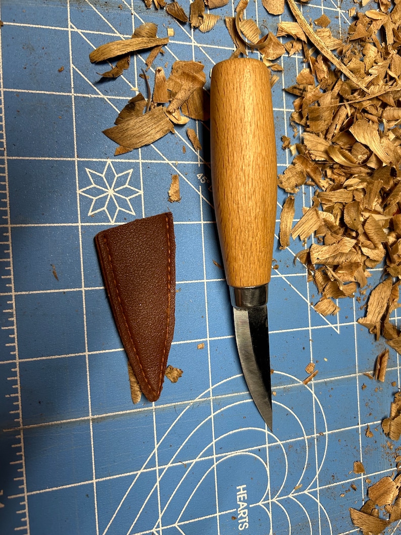 May include: A small, wooden-handled carving knife with a curved blade, resting on a blue cutting mat with wood shavings. A brown leather sheath is next to the knife.