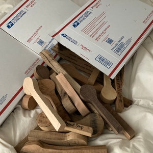 May include: A collection of unfinished wooden spoons in various shapes and sizes, alongside several USPS Priority Mail boxes. The spoons are made from light and dark wood, some with text. The boxes are white with red and blue accents.