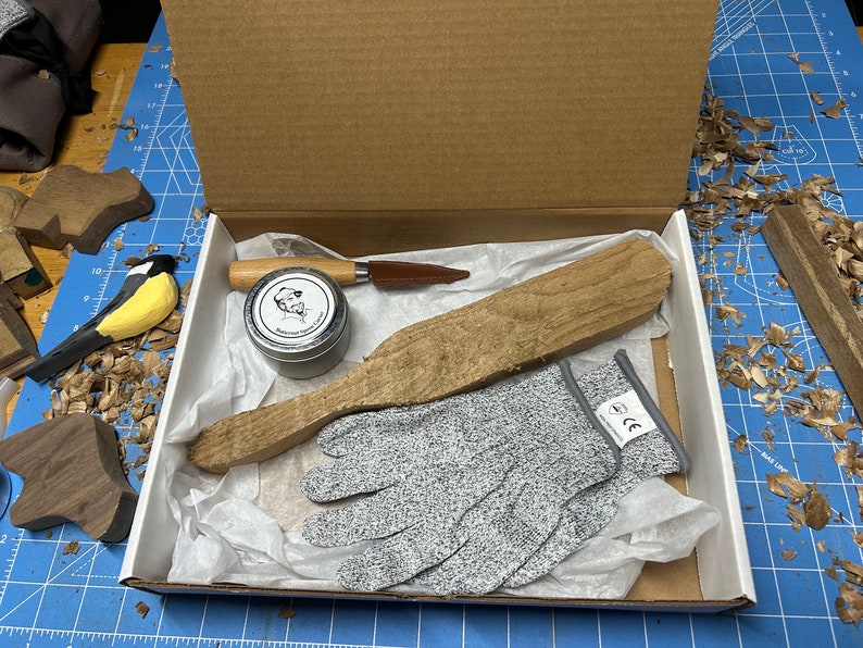 May include: A wooden box filled with woodworking tools and supplies. The box contains a wooden paddle, a small tin of wood conditioner, a small knife, and a pair of gray safety gloves.