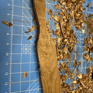 May include: A wooden paddle with a rounded top and a flat bottom, resting on a blue cutting mat with wood shavings scattered around it. The paddle is a light brown color and has a smooth surface.