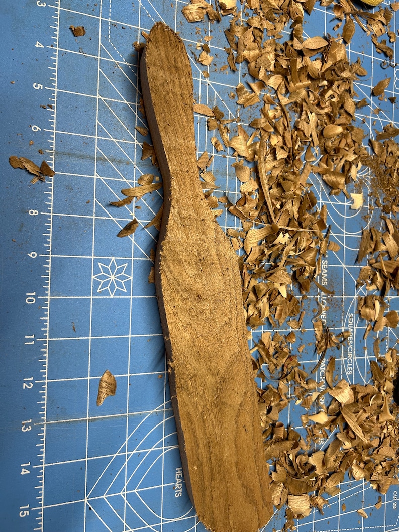 May include: A wooden paddle with a rounded top and a flat bottom, resting on a blue cutting mat with wood shavings scattered around it. The paddle is a light brown color and has a smooth surface.