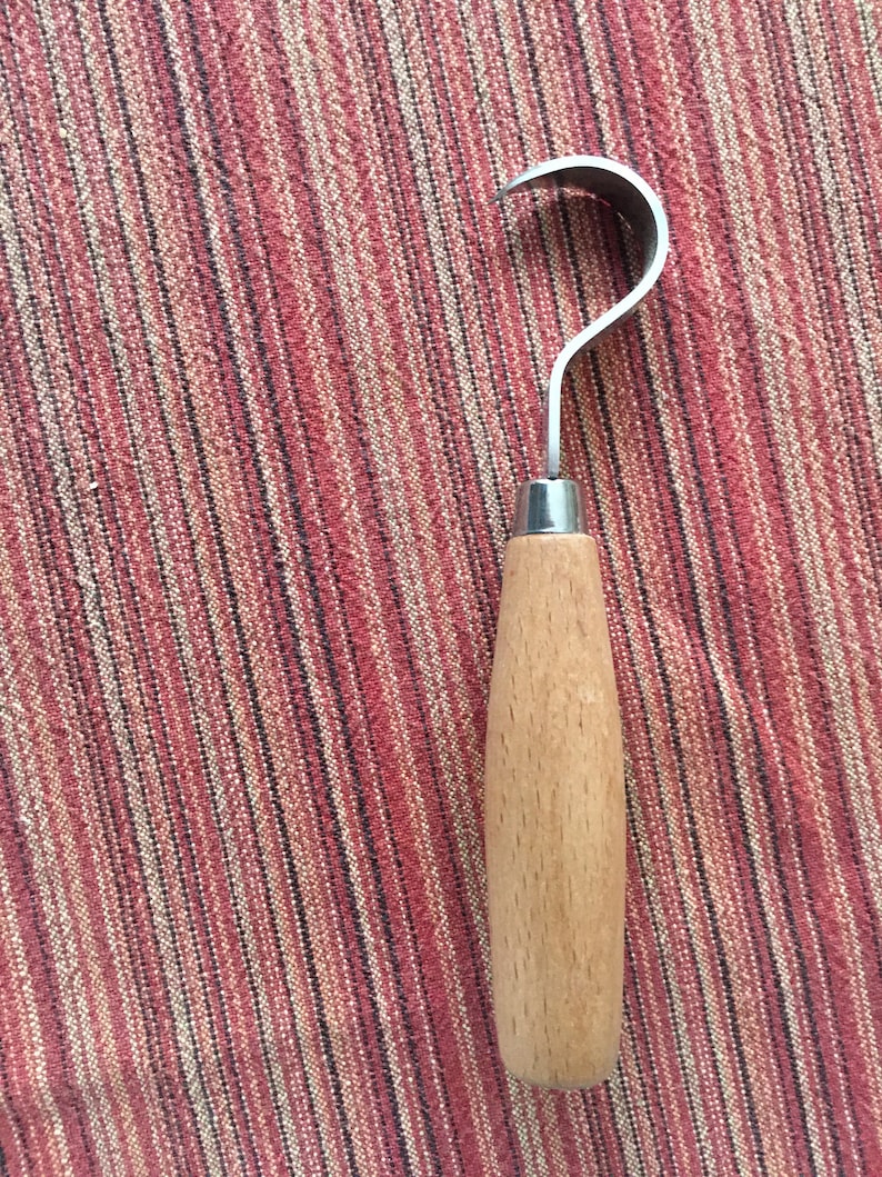 May include: A wooden handled carving tool with a curved metal blade. The tool is on a striped red and white fabric background.