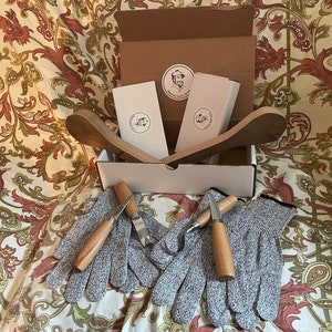K&ouml;nnte beinhalten: Eine Schachtel mit zwei Holzl&ouml;ffeln, zwei Paar grauen Schutzhandschuhen und zwei kleinen Werkzeugen mit Holzgriffen. Die Schachtel ist ge&ouml;ffnet und die Gegenst&auml;nde sind sichtbar. Die Schachtel steht auf einem gemusterten Stoff.