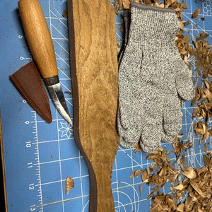 May include: A close-up of a wooden paddle with a handle, a brown leather knife sheath, a wooden-handled knife, and a pair of gray safety gloves. The items are on a blue cutting mat with white grid lines. There are wood shavings scattered around the items.