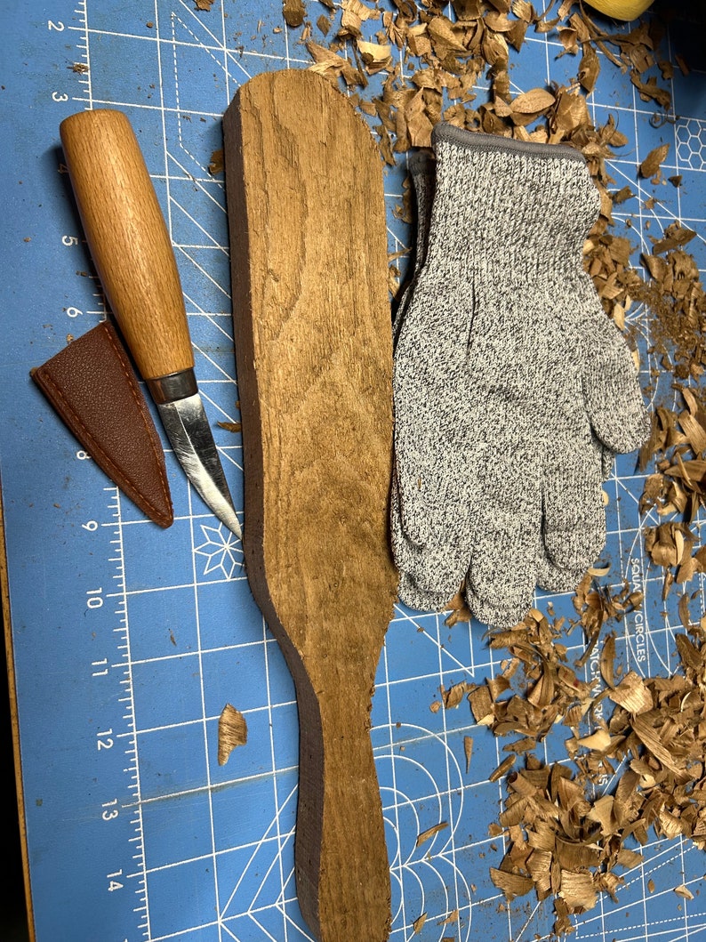 May include: A close-up of a wooden paddle with a handle, a brown leather knife sheath, a wooden-handled knife, and a pair of gray safety gloves. The items are on a blue cutting mat with white grid lines. There are wood shavings scattered around the items.