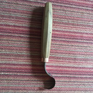 May include: A wooden handled carving tool with a curved metal blade. The tool is on a red and white striped surface.
