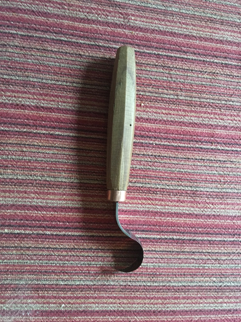 May include: A wooden handled carving tool with a curved metal blade. The tool is on a red and white striped surface.