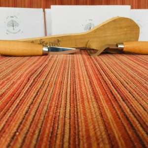 May include: Two wood carving tools with wooden handles on a striped orange and brown surface. The tools are a knife and a hook.