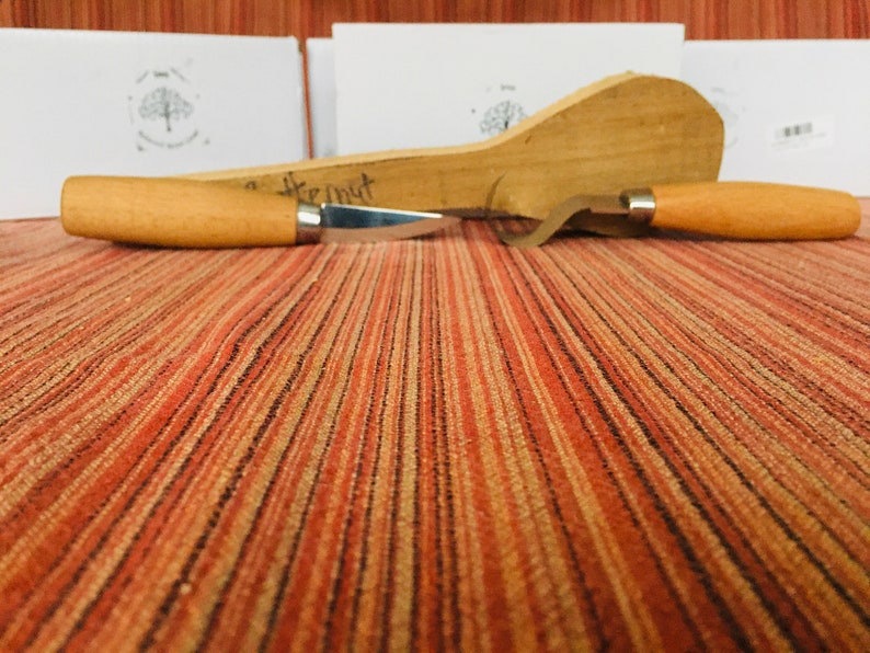 May include: Two wood carving tools with wooden handles on a striped orange and brown surface. The tools are a knife and a hook.