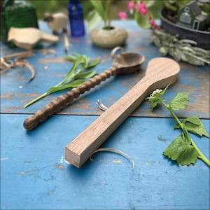K&ouml;nnte beinhalten: Zwei Holzl&ouml;ffel auf einer blauen Holzoberfl&auml;che. Ein L&ouml;ffel ist mit einem dekorativen Muster geschnitzt, der andere ist schlicht. Beide L&ouml;ffel sind aus hellem braunem Holz.