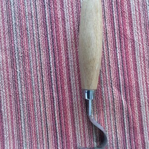 May include: A wooden handled carving tool with a curved metal blade. The tool is on a striped fabric background.