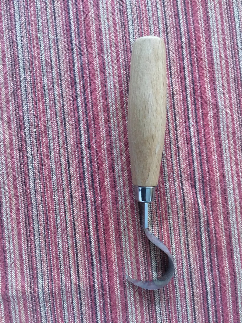 May include: A wooden handled carving tool with a curved metal blade. The tool is on a striped fabric background.