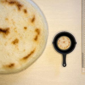 May include: A large, round, golden-brown arepa next to a miniature black frying pan containing a smaller arepa. A ruler shows the size of the arepa. The arepa is a traditional Latin American food.