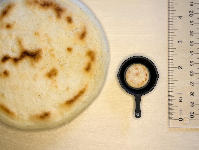 May include: A large, round, golden-brown arepa next to a miniature black frying pan containing a smaller arepa. A ruler shows the size of the arepa. The arepa is a traditional Latin American food.