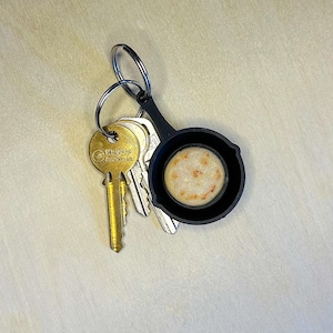 May include: A set of keys with a gold-colored key and a black keychain shaped like a miniature frying pan. The pan holds a small, round, beige object with brown spots. The keys are on a light-colored surface.