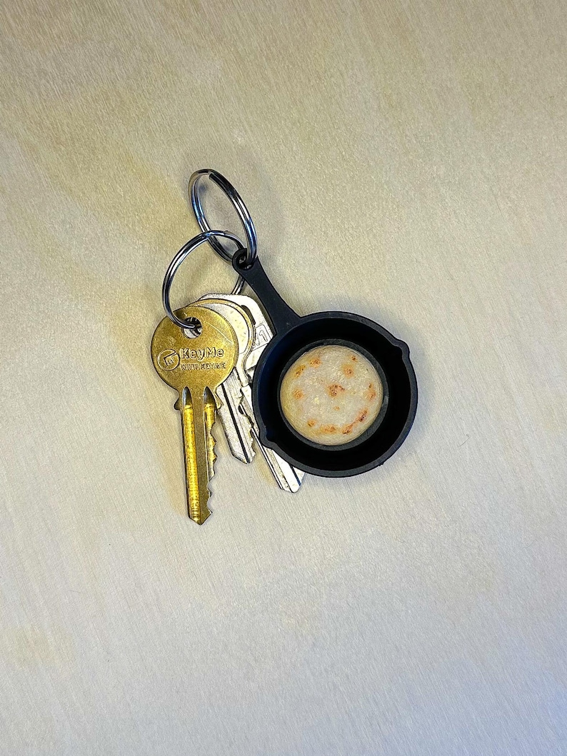 May include: A set of keys with a gold-colored key and a black keychain shaped like a miniature frying pan. The pan holds a small, round, beige object with brown spots. The keys are on a light-colored surface.