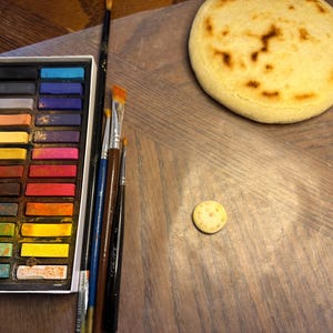 May include: A still-life composition featuring a palette of colorful pastels, various paintbrushes, a round, golden-brown baked item, and a small, round cracker. The items are arranged on a wooden surface, suggesting an art or culinary theme.