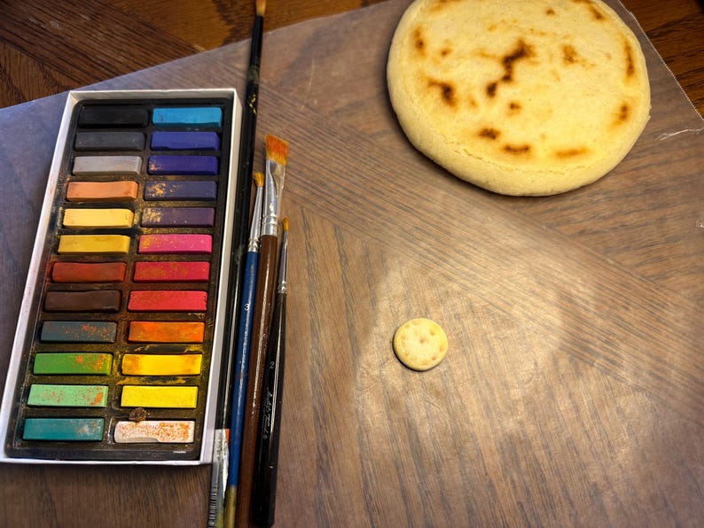 May include: A still-life composition featuring a palette of colorful pastels, various paintbrushes, a round, golden-brown baked item, and a small, round cracker. The items are arranged on a wooden surface, suggesting an art or culinary theme.