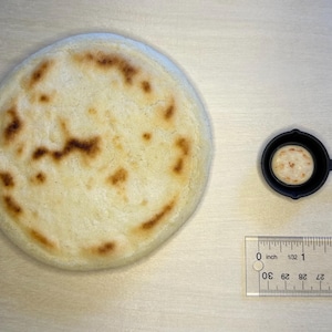 May include: A large, round, lightly browned arepa next to a miniature cast iron skillet with a smaller arepa inside. A ruler shows the large arepa is approximately 3 inches in diameter. The items are on a light wood surface.