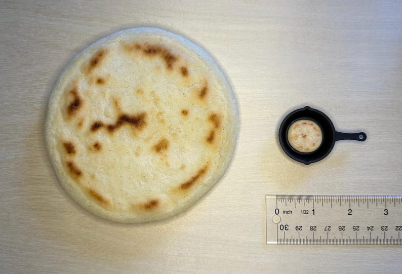 May include: A large, round, lightly browned arepa next to a miniature cast iron skillet with a smaller arepa inside. A ruler shows the large arepa is approximately 3 inches in diameter. The items are on a light wood surface.