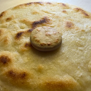 May include: A close-up shot of a round, golden-brown arepa, a type of cornmeal cake, with a smaller, circular piece on top. The arepa has dark brown spots from being cooked. The background is a light brown surface.