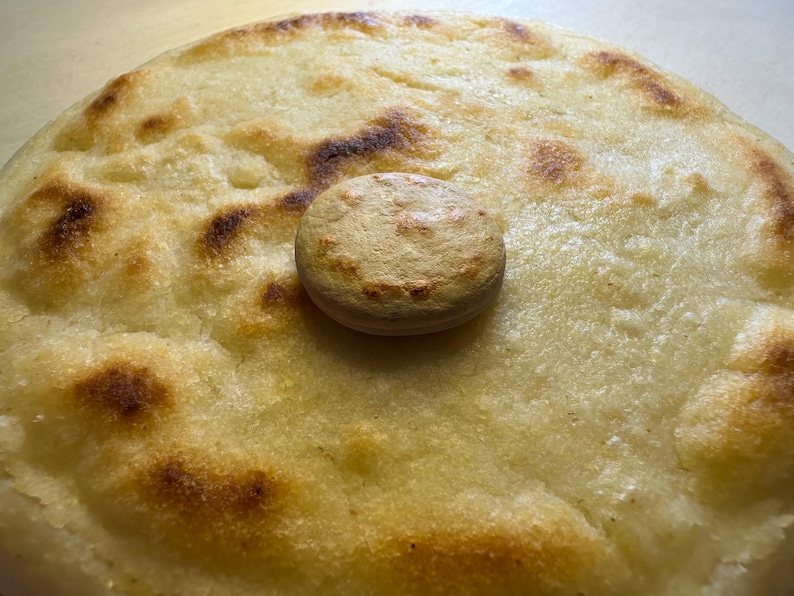 May include: A close-up shot of a round, golden-brown arepa, a type of cornmeal cake, with a smaller, circular piece on top. The arepa has dark brown spots from being cooked. The background is a light brown surface.