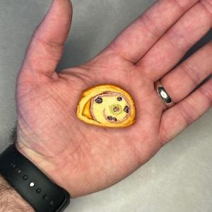 Puede incluir: Un pequeño y detallado rollo de pastelería con un diseño en espiral. La pastelería tiene un exterior marrón dorado y un relleno con manchas oscuras y un centro verde. El artículo se sostiene en la mano de una persona.