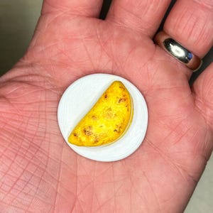 Peut inclure: Une maquette miniature et détaillée d'une empanada brun doré sur une petite assiette blanche. L'empanada a une surface texturée avec des taches plus foncées, suggérant une apparence cuite. L'assiette est ronde et unie.