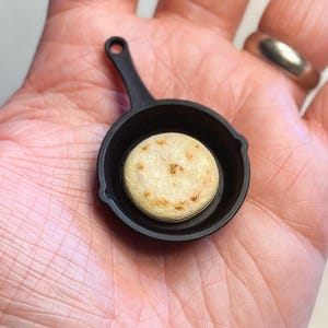 May include: A miniature black cast iron skillet holding a small, round, golden-brown arepa. The arepa has a slightly textured surface with darker spots. The skillet has a small handle and is set against a neutral background.