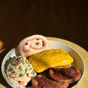 May include: A white ceramic plate holds several food-shaped sculptures. The plate contains a roll with a pink swirl and green olive, a yellow rectangular item, a salad with green peas and carrots, and two brown, round items.